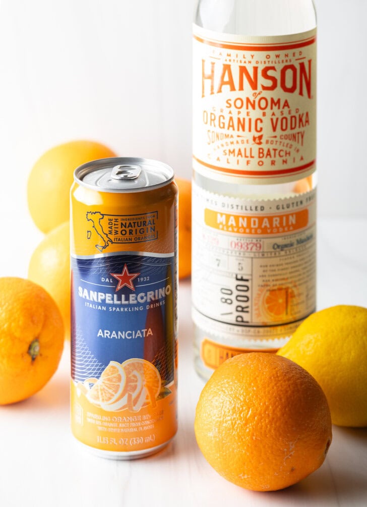 Can of orange soda, bottle of vodka, several fresh oranges and lemons all around.