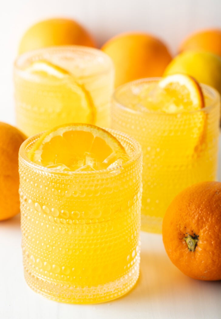 Two orange crush drinks in glasses, each garnished with a slice of orange. There are fresh oranges and lemons around the glasses.