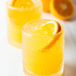 Two orange crush cocktails in glasses, each garnished with a slice of orange. There are fresh oranges and lemons around the glasses.