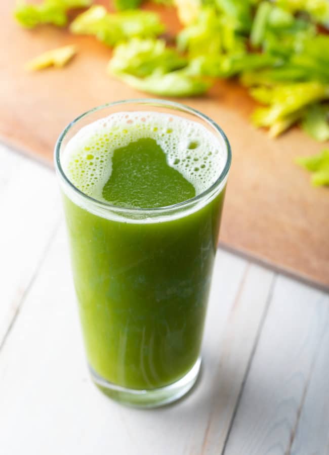 How To Make Celery Juice - Large glass of celery juice with celery on a cutting board in the background.