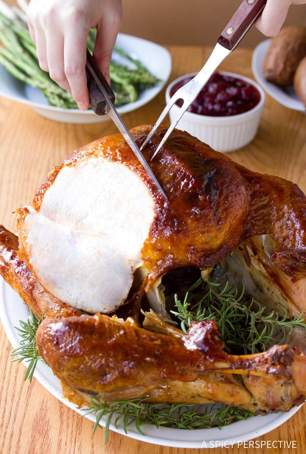 Hands carving into a whole roast turkey on a serving platter lined with herbs. 