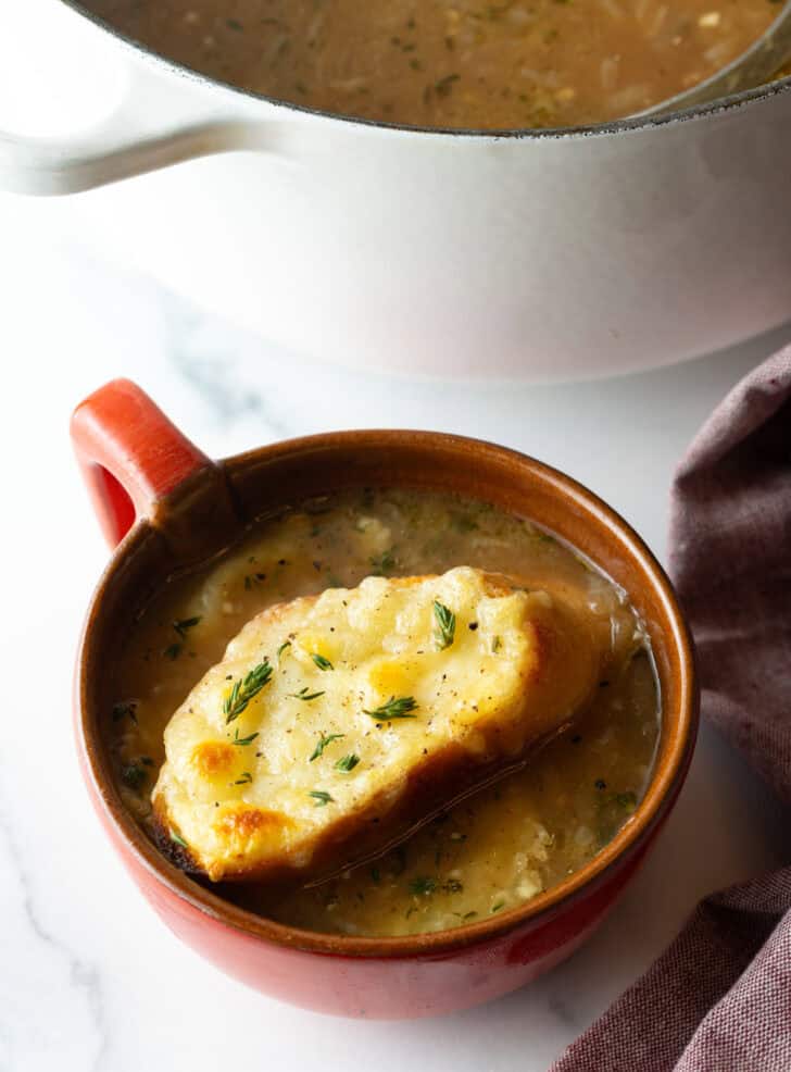 Large red mug with French onion soup, topped with a slice of baguette with melted cheese.