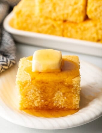 Square of cornbread is on a white plate, topped with a pat of butter and drizzled with honey.