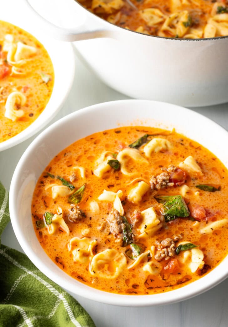 Top down view white bowl with creamy sausage and tortellini soup. The large pot of soup is off to the side.